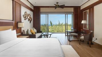 A hotel room with a large bed, a sofa, a small table, and a chair. There is a balcony with a view of the ocean.