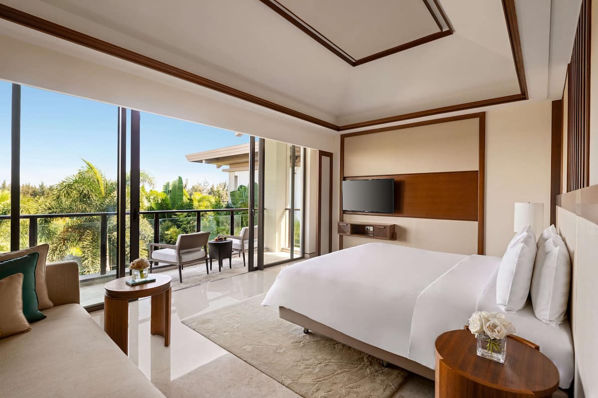 A modern hotel room with a large bed, a TV, and a balcony with a view of greenery.
