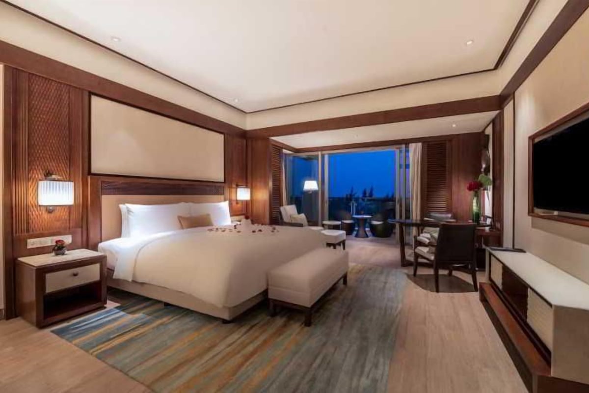 A modern hotel room with a large bed, a desk, a TV, and a balcony with a view.