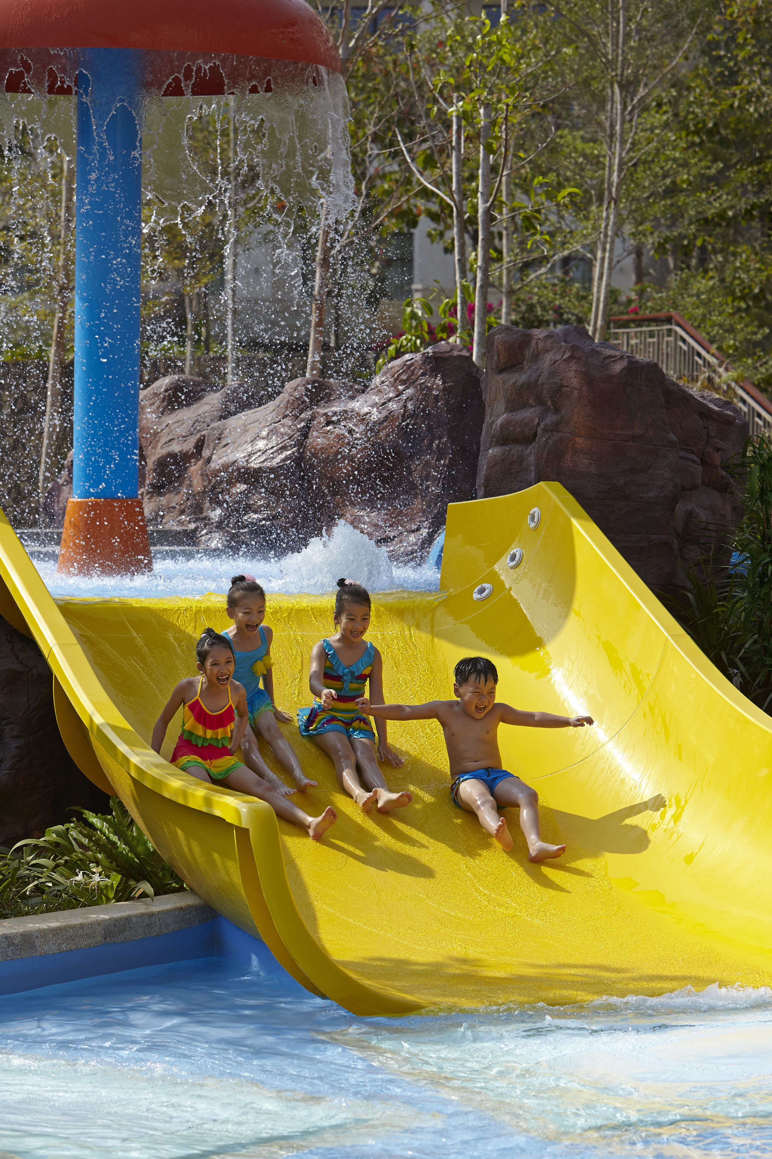 Un toboggan aquatique avec trois enfants en haut, une tour d’eau bleue et rouge, et un paysage rocheux en arrière-plan.