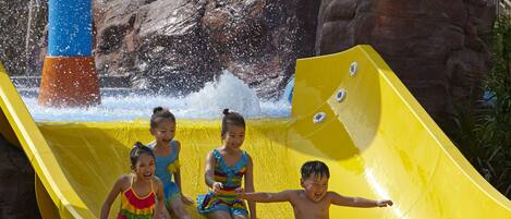 Un toboggan aquatique avec trois enfants en haut, une tour d’eau bleue et rouge, et un paysage rocheux en arrière-plan.