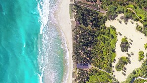Vista aérea de una zona costera con playa, vegetación densa y un edificio pequeño.