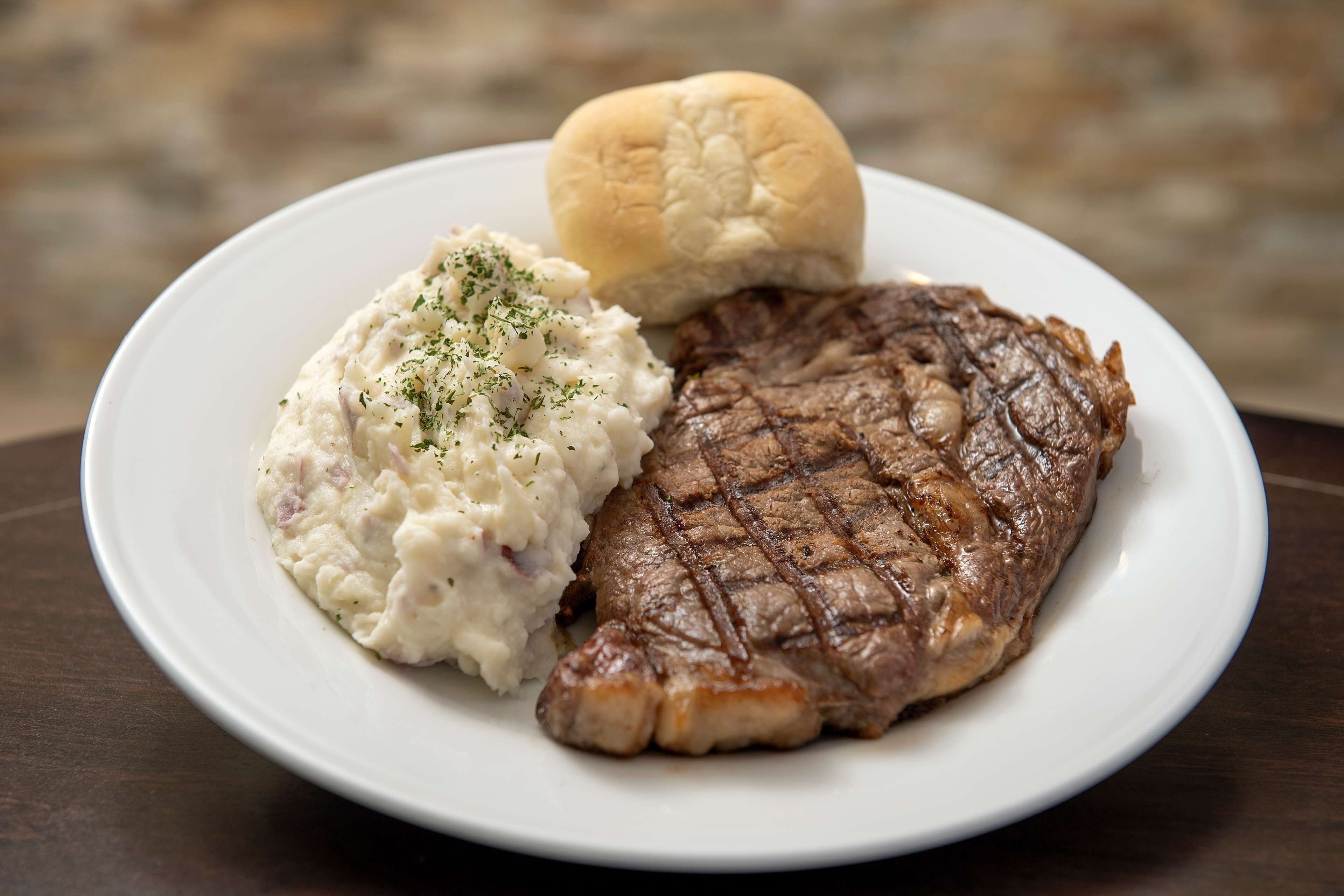 Une assiette comprenant un steak grillé, de la purée de pommes de terre et un petit pain.