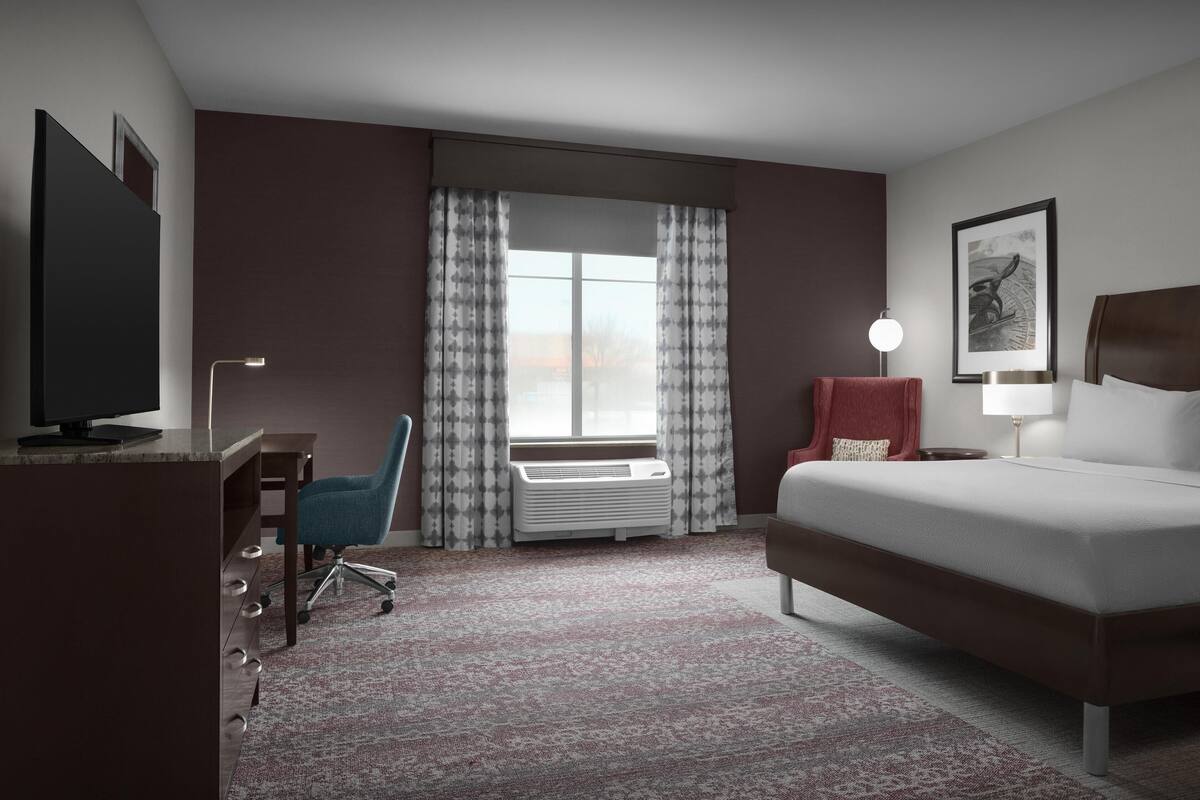 Habitación de hotel con cama, escritorio, silla, aire acondicionado, ventana con cortinas y lámpara.
