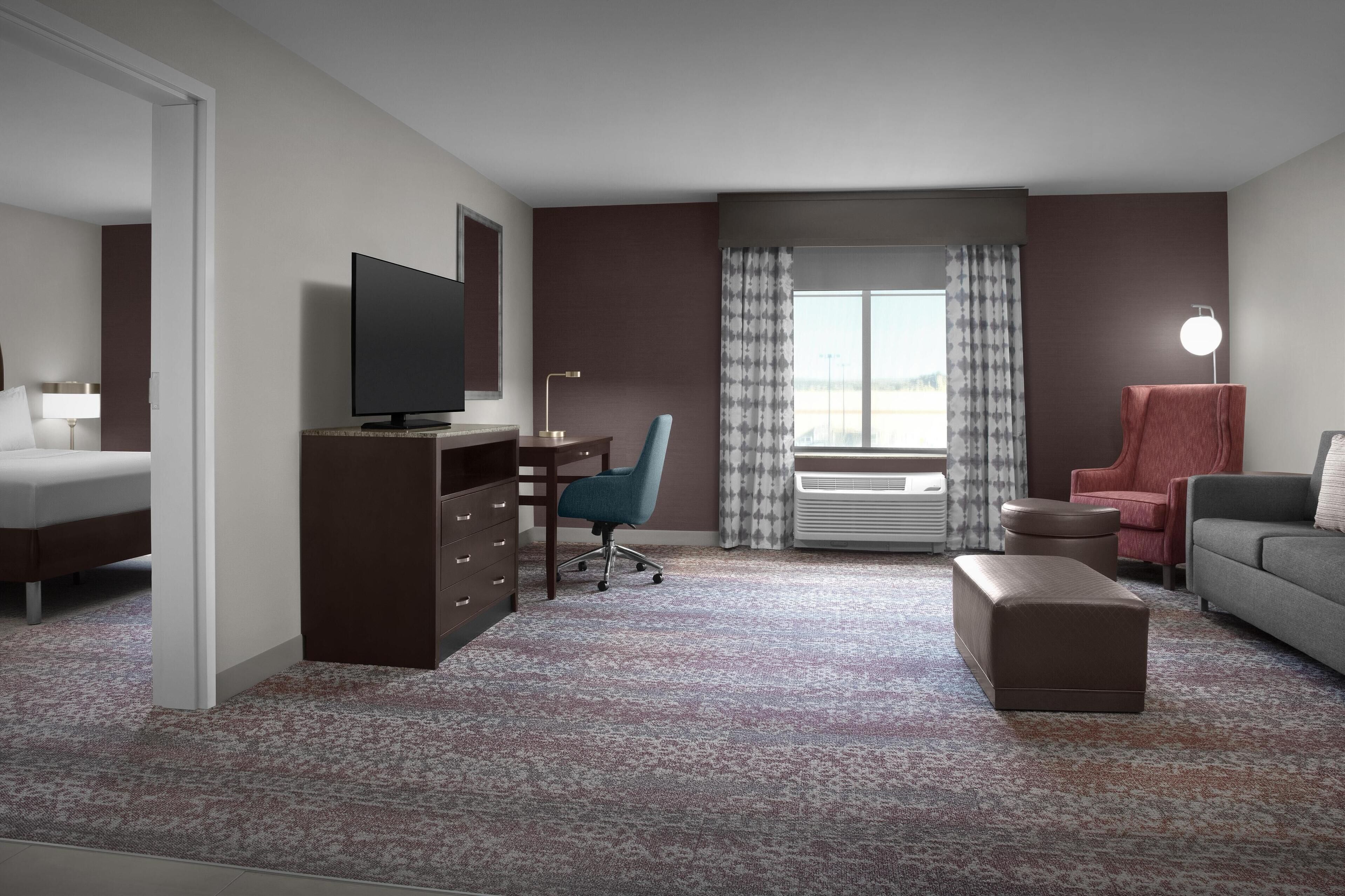 A hotel room with a bed, a desk with a chair, a television, a sofa, a red chair, a lamp, and a large window with curtains.