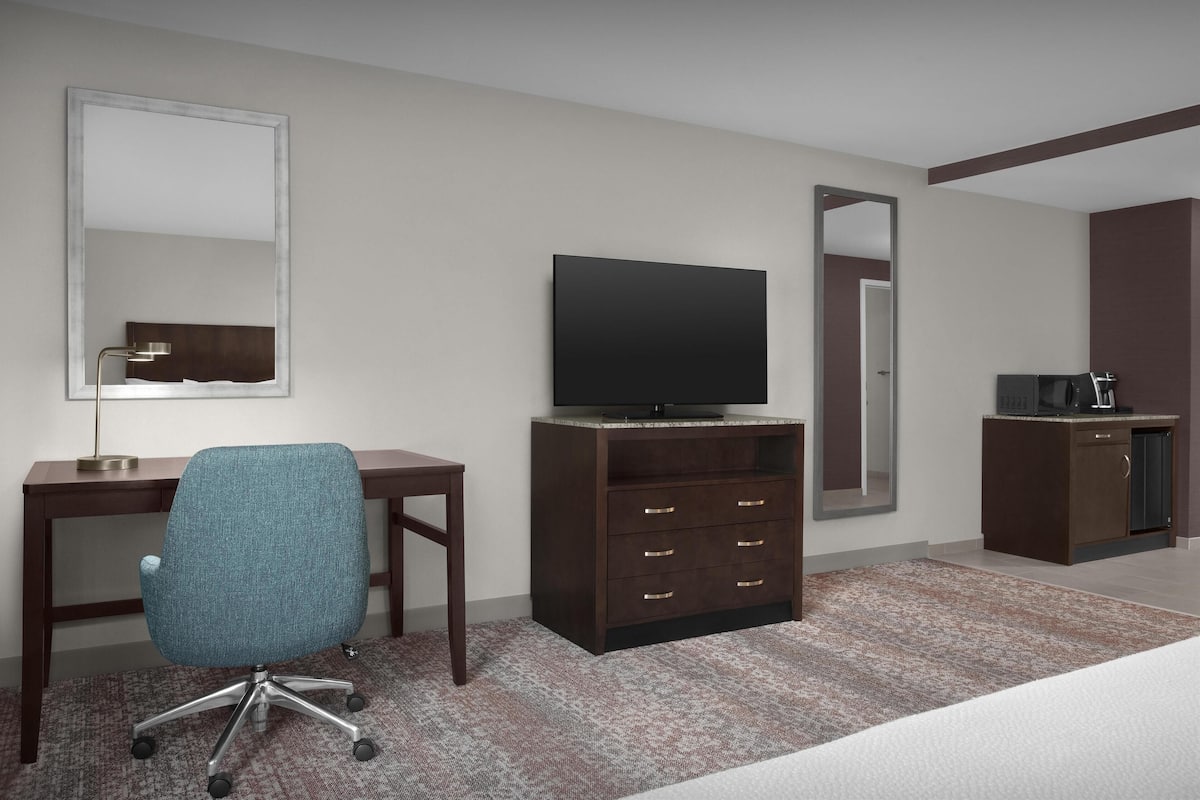 Una habitación de hotel con escritorio, silla, televisor, armario y una pequeña kitchenette.