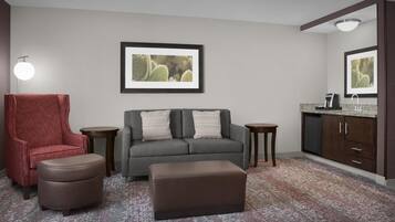 Una sala de estar con un sofá gris, un sillón rojo, un reposapiés marrón y una pequeña mesa. Hay un cuadro enmarcado en la pared y una zona de cocina con armarios y una encimera.