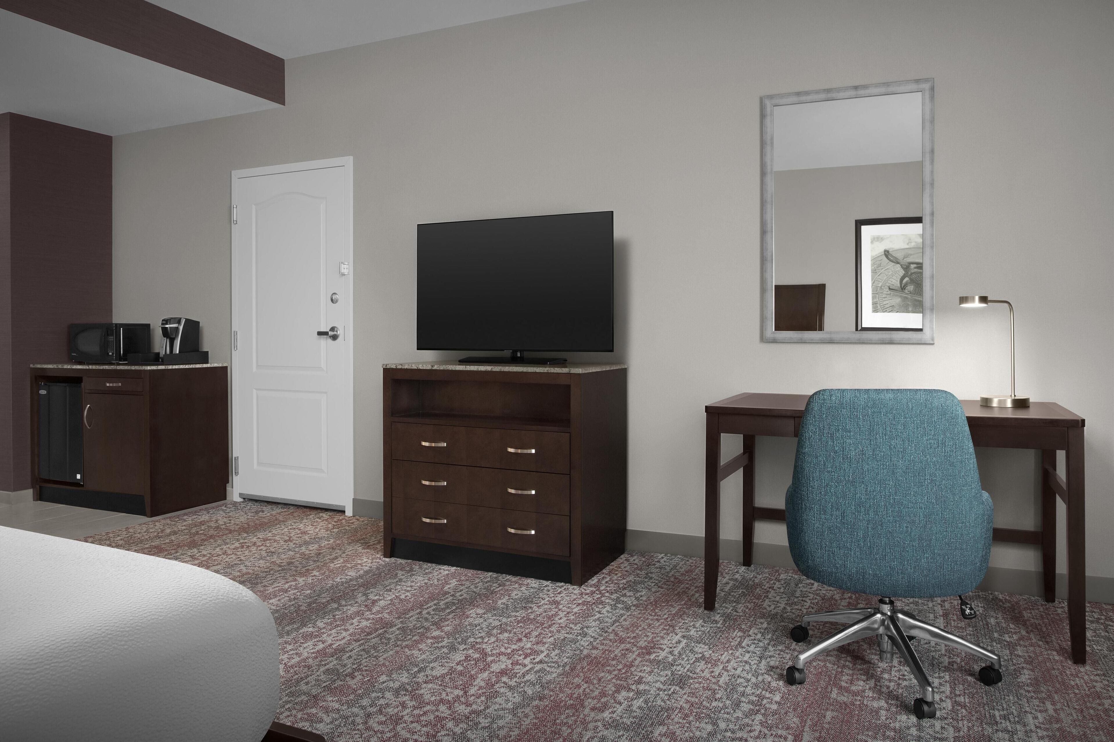 Habitación de hotel con cama, televisor sobre un mueble de madera, escritorio con silla, lámpara de escritorio y espejo en la pared.