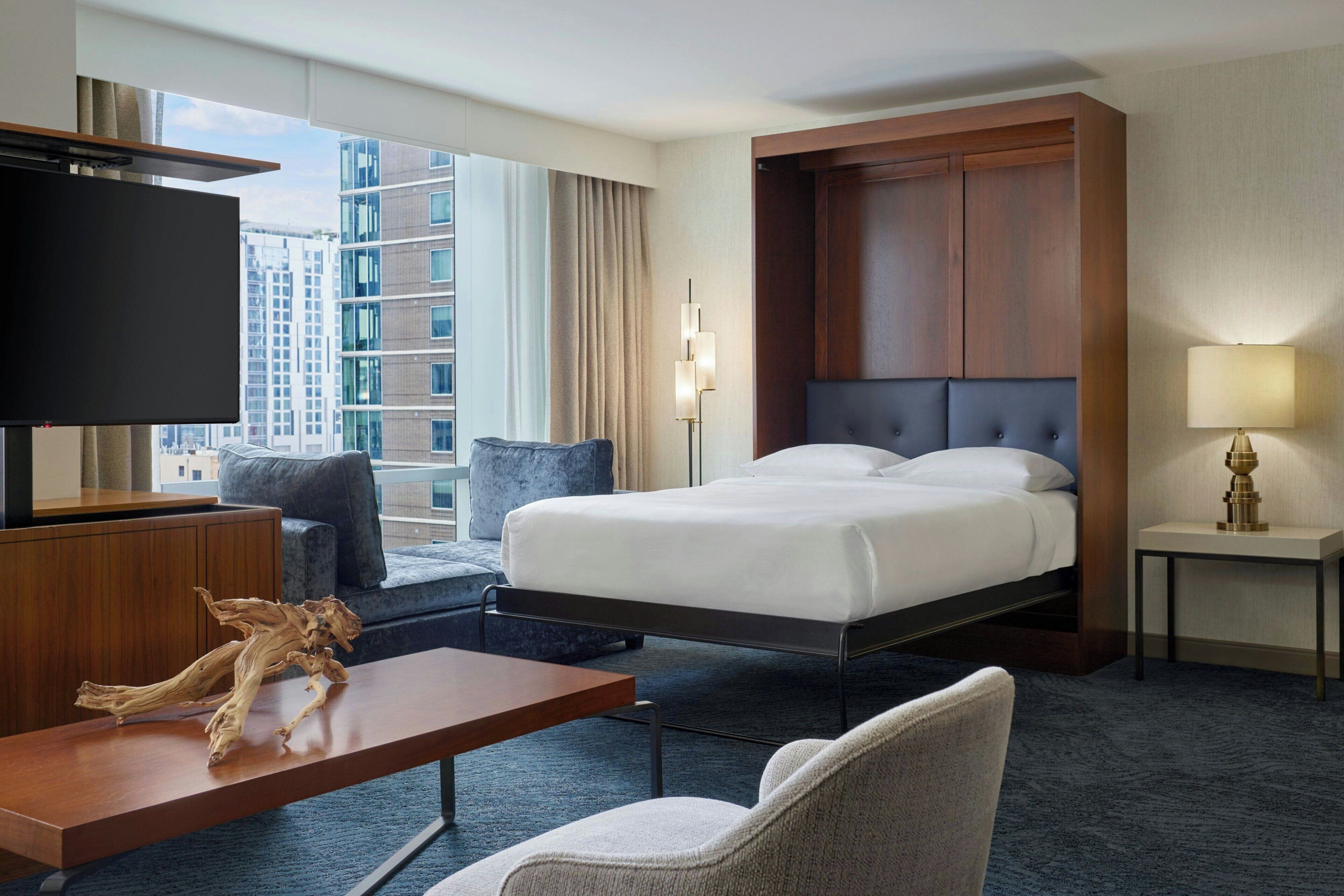 Habitación de hotel con cama, televisor, escritorio, silla, lámpara y vista a la ciudad.