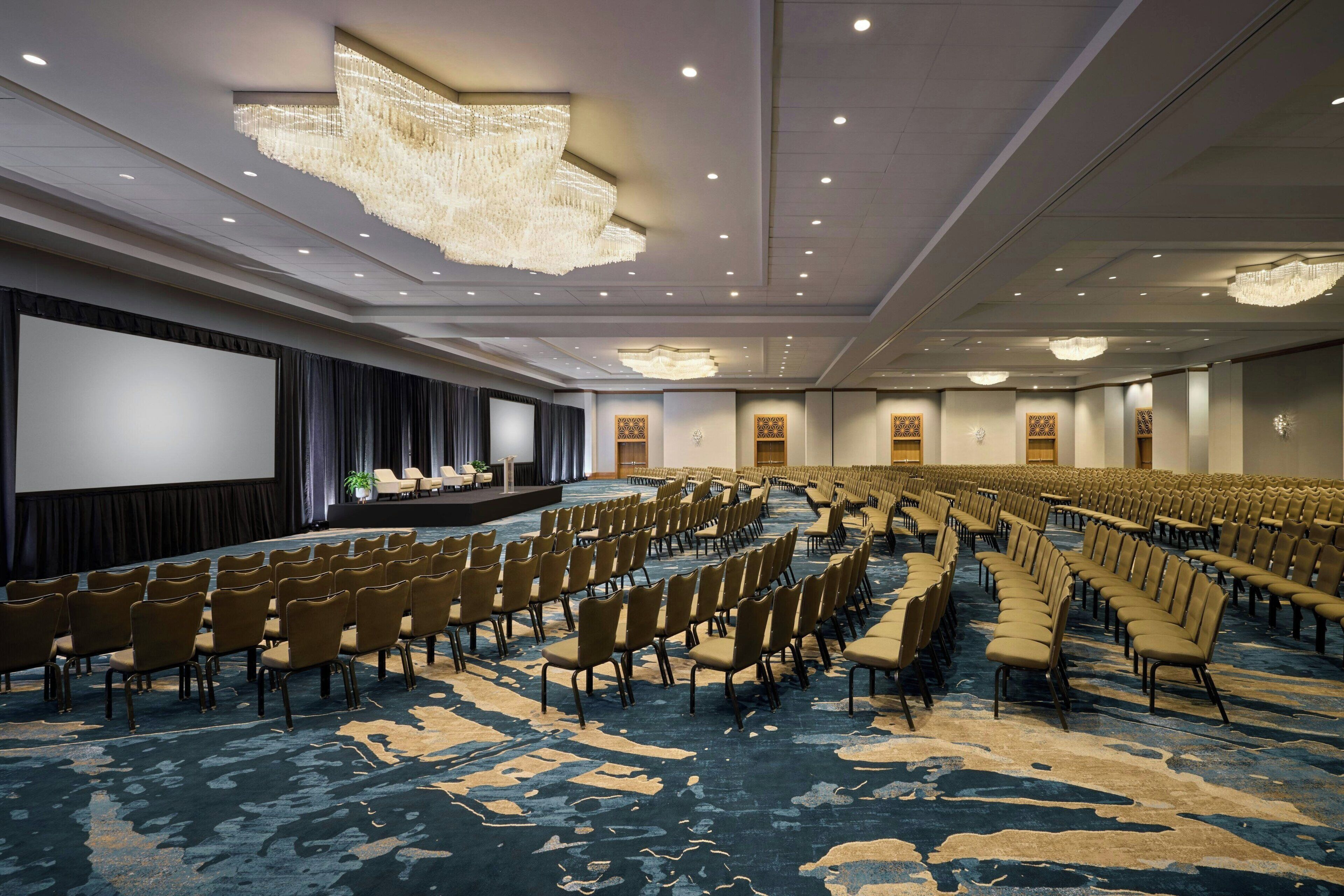 Een ruime congreszaal met rijen stoelen, een podium en een groot scherm.