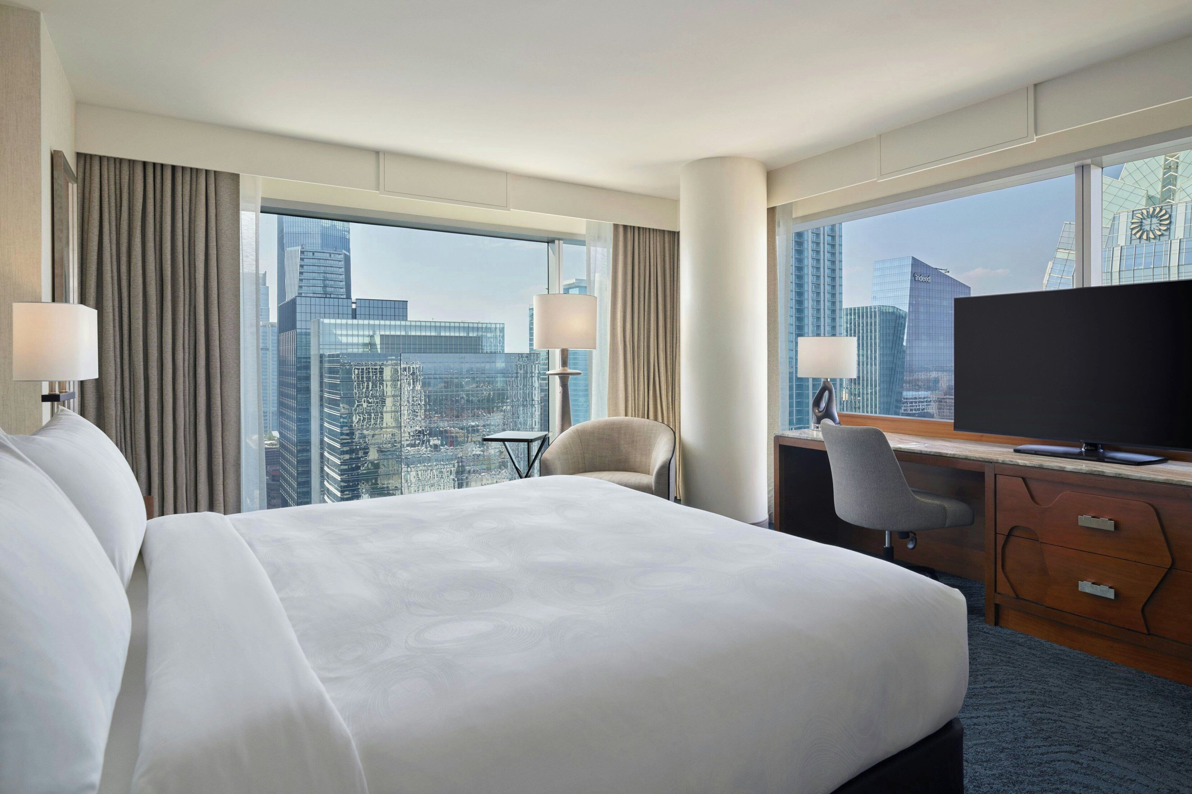A hotel room with a large bed, a desk with a computer, a chair, and a TV. The room has large windows with a city view.