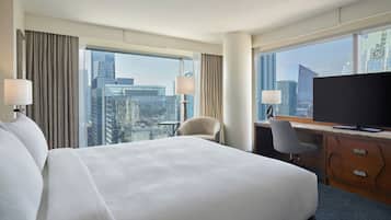 A hotel room with a large bed, a desk with a computer, a chair, and a TV. The room has large windows with a city view.