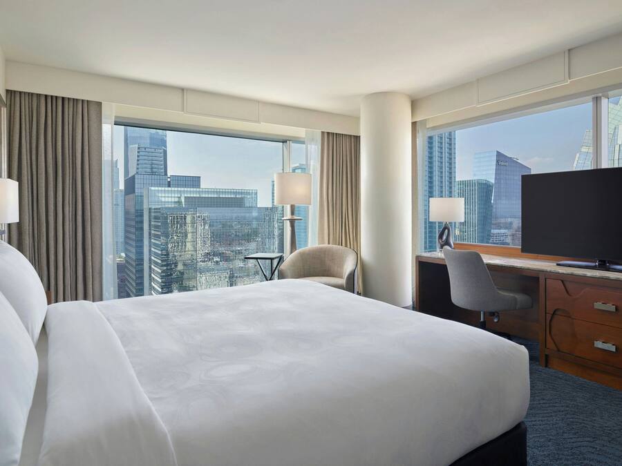 A hotel room with a large bed, a desk with a computer, a chair, and a TV. The room has large windows with a city view.