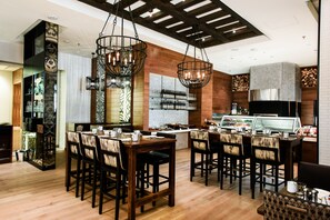 A restaurant with wooden flooring, a central dining table with chairs, hanging metal chandeliers, and a counter with a display case. - JW Marriott Austin (Austin)