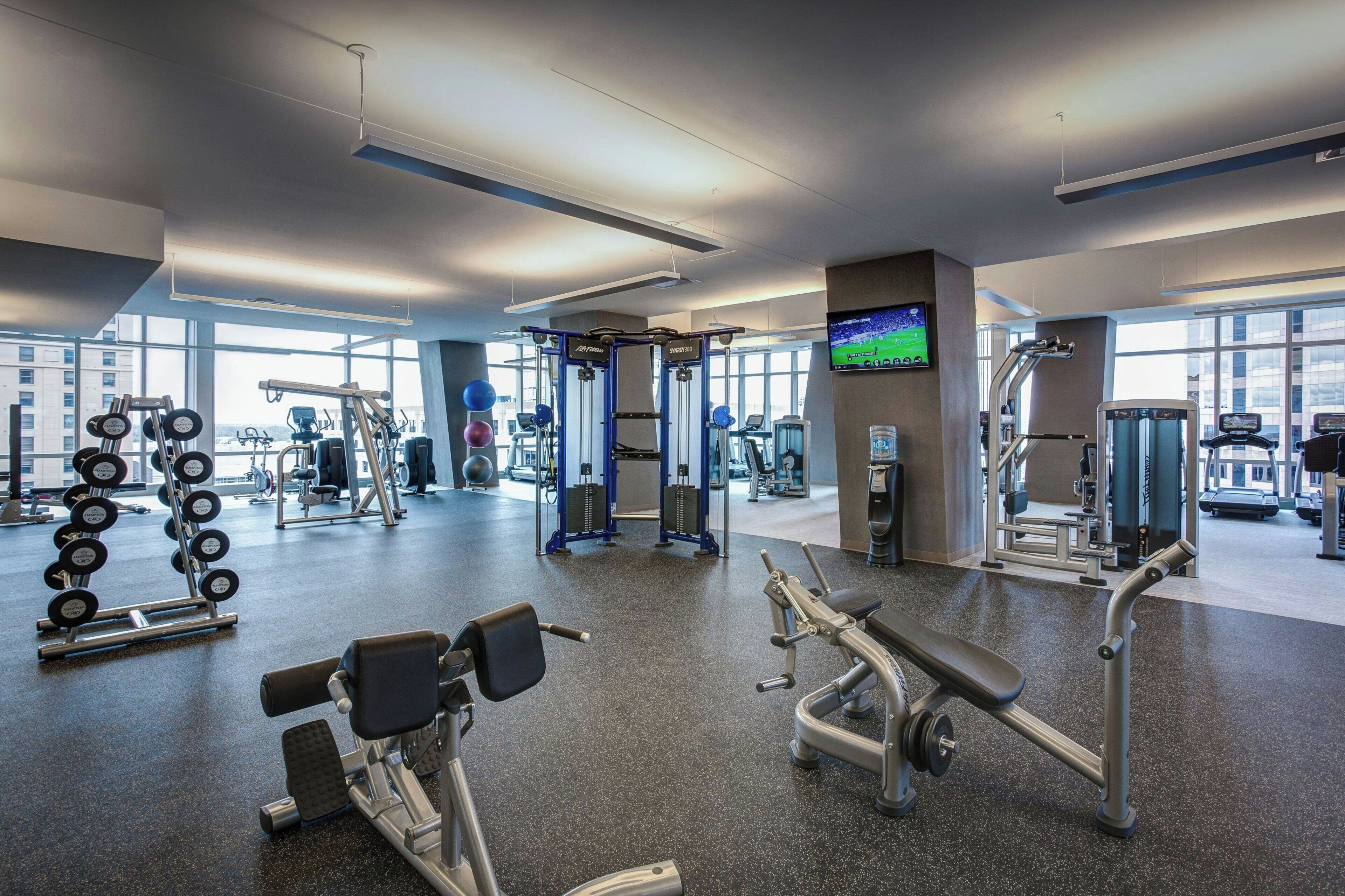 A modern gym with various exercise equipment, large windows, and a television screen.