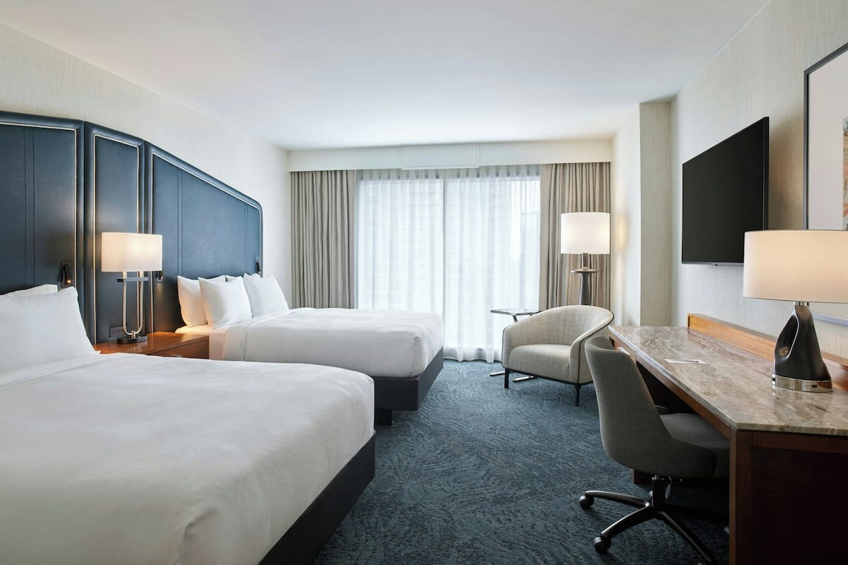 Ein Hotelzimmer mit zwei Betten, einem Schreibtisch, einem Stuhl, einem Fernseher und einem großen Fenster mit Vorhängen.