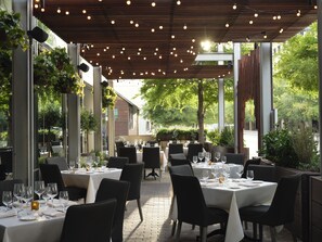 An outdoor dining area with tables set for guests, surrounded by greenery and a wooden pergola with string lights. - JW Marriott Austin (Austin)