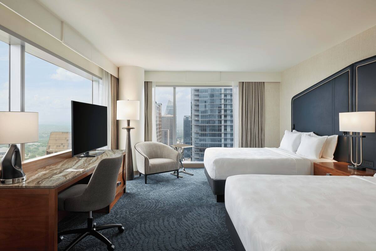 Ein Hotelzimmer mit zwei Betten, einem Schreibtisch, einem Sessel, einem Fernseher und großen Fenstern mit Blick auf die Stadt.
