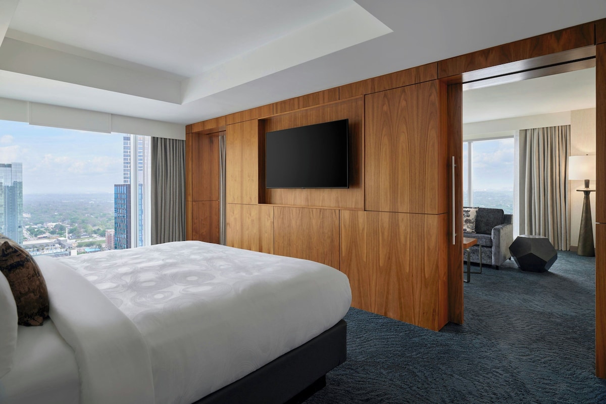 A modern hotel room with a large bed, a flat-screen TV mounted on the wall, and a city view.