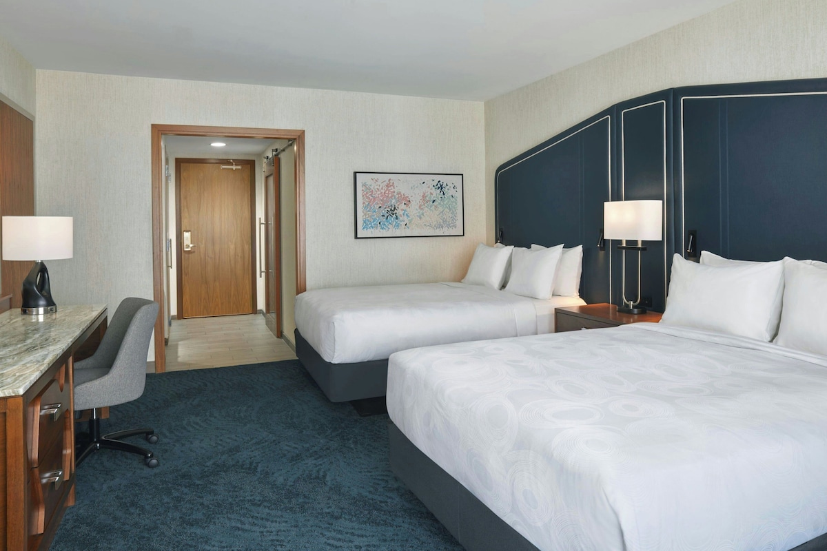 A hotel room with two beds, a desk with a chair, a desk lamp, and a door leading to another room.