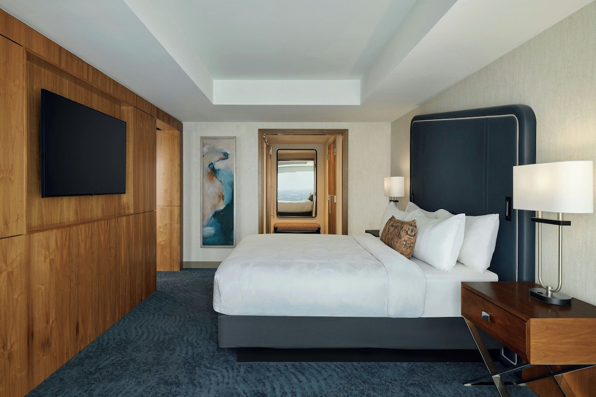 A hotel room with a large bed, a TV mounted on the wall, a painting, and a view through an open door.
