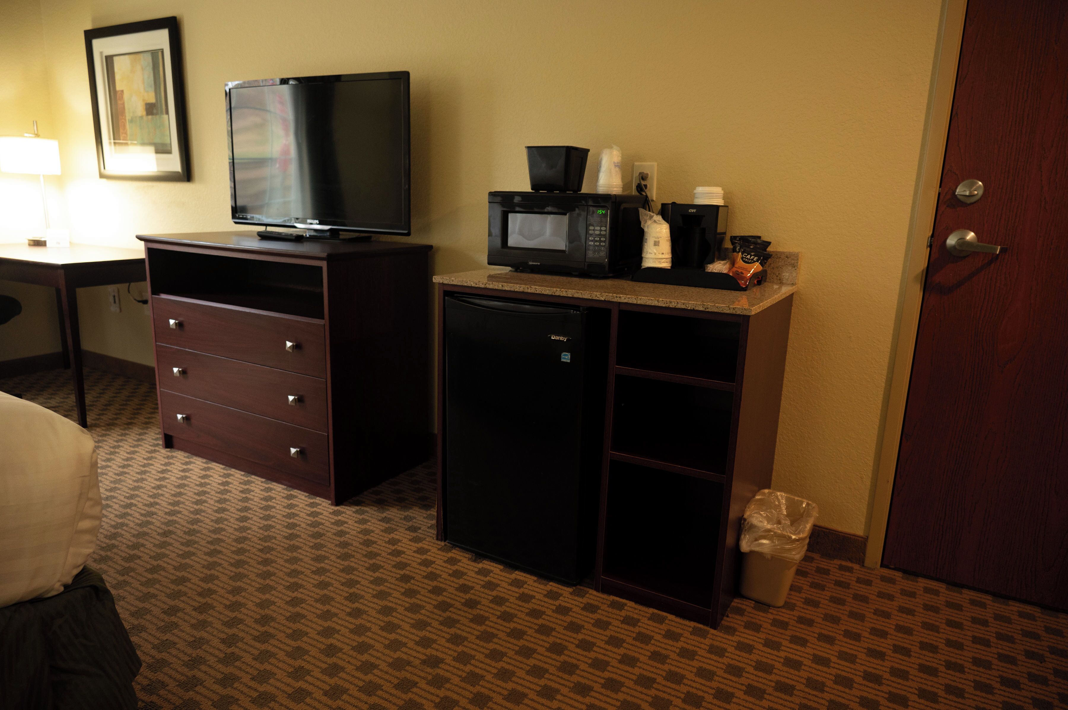 A hotel room with a television, a microwave, a coffee maker, a trash bin, and a desk with a lamp.