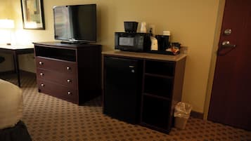 A hotel room with a television, a microwave, a coffee maker, a trash bin, and a desk with a lamp.