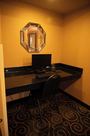 Habitación de hotel con escritorio, computadora, silla, espejo y alfombra estampada.