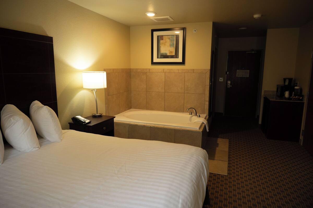 Ein Hotelzimmer mit einem großen Bett, einem Nachttisch mit Lampe, einem Badezimmer mit Badewanne und einem gerahmten Bild an der Wand.