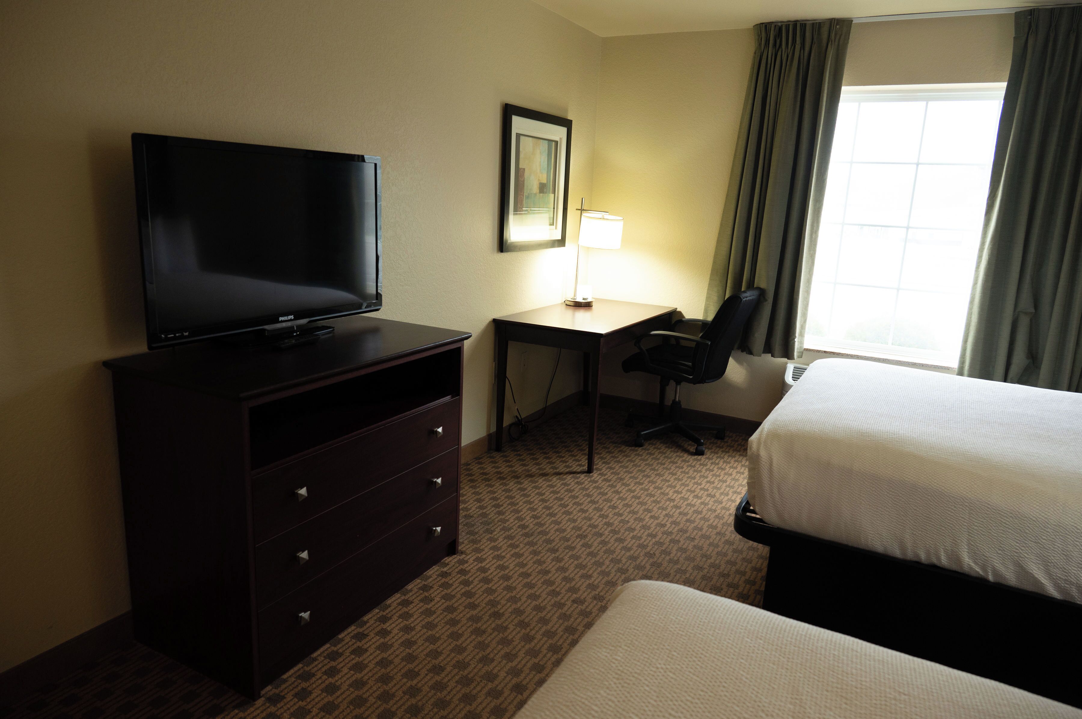 Habitación de hotel con televisor sobre una cómoda, un escritorio con silla, una cama y una ventana con cortinas.