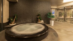 Zona de spa con bañera de hidromasaje, paredes de textura oscura y plantas en macetas.
