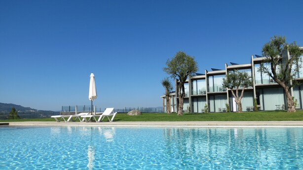 Una piscina con sillones de descanso y una sombrilla, un edificio moderno con balcones, y un cielo azul despejado.