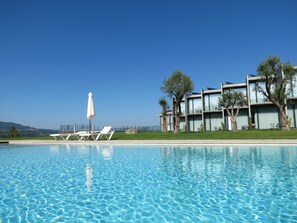 En swimmingpool med liggestole og en parasol, en moderne bygning med altaner, og en klar blå himmel.
