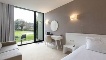 A hotel room with a bed, a desk, a chair, a round mirror, and a view of a garden through a large window.