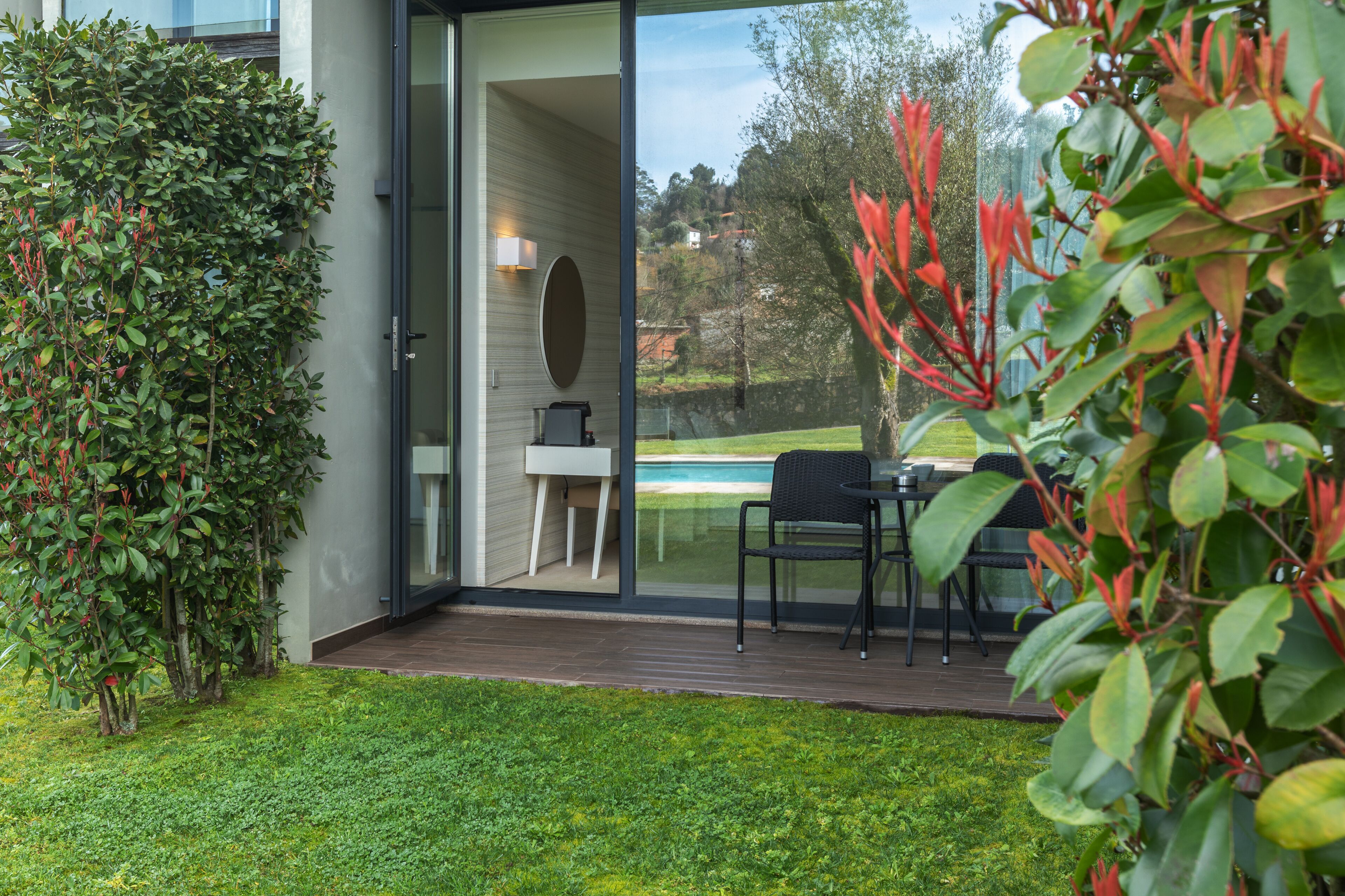 Un patio moderno al aire libre con puerta de vidrio que da a un comedor, rodeado de abundante vegetación y vistas a una piscina.