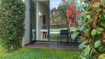 A modern outdoor patio with a glass door leading to a dining area, surrounded by lush greenery and a view of a swimming pool.