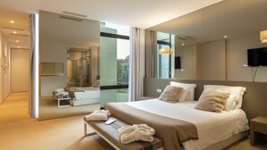 Una habitación de hotel moderna con una cama grande, un banco con toallas, un baño con ducha de vidrio y un televisor montado en la pared.