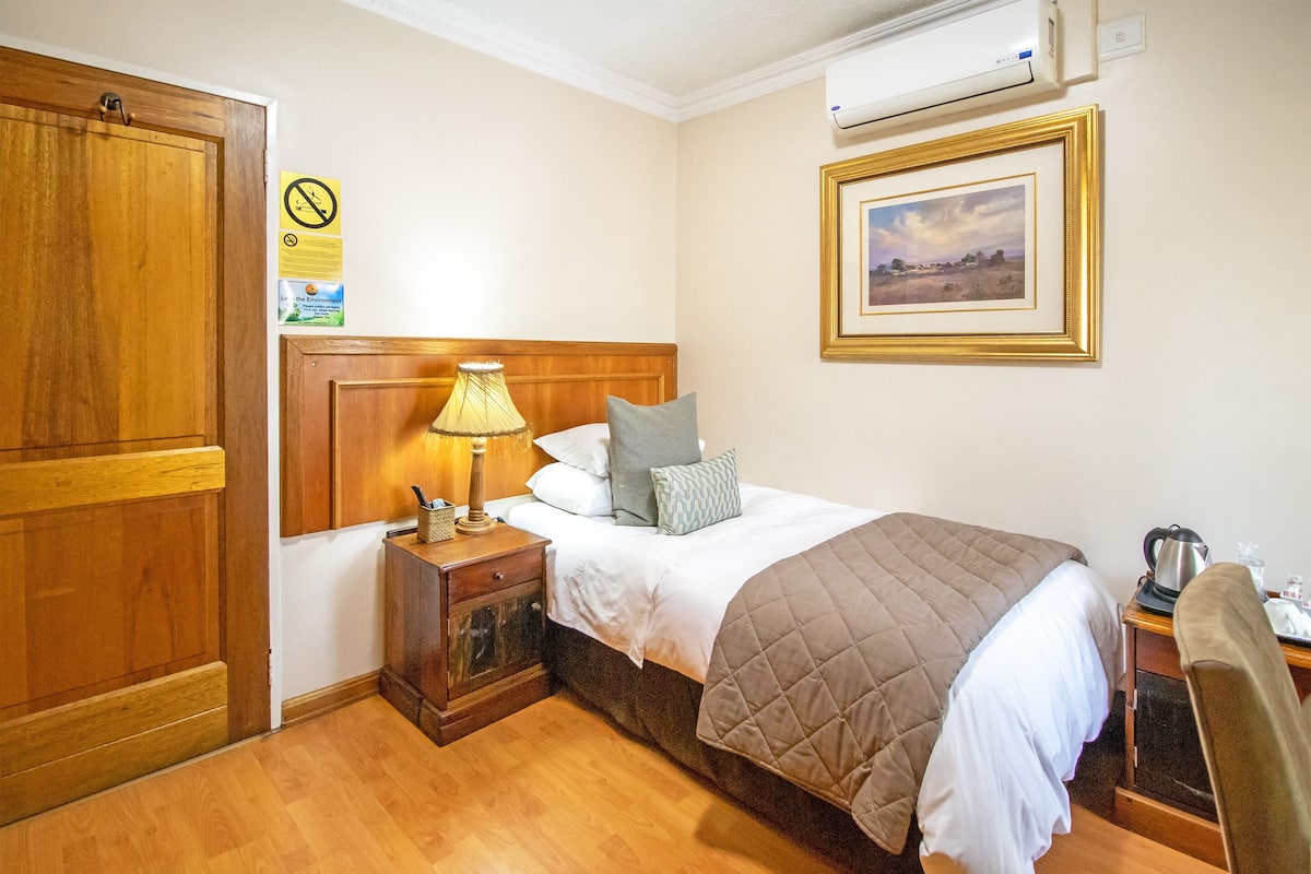 A hotel room with a wooden wardrobe, a bedside table with a lamp, a bed with a quilted blanket, a framed picture on the wall, and a coffee maker on the desk.