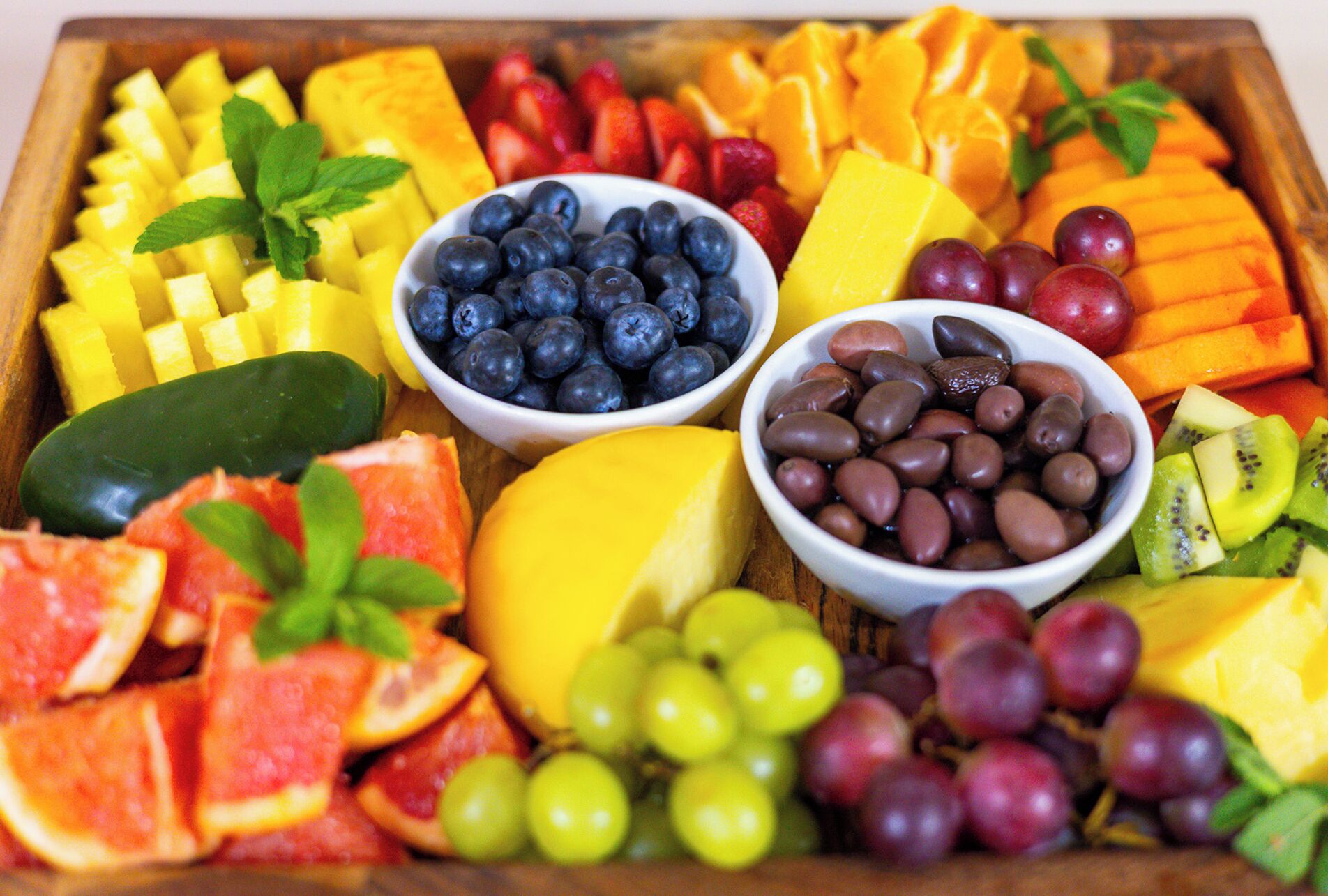A wooden tray filled with various fruits and vegetables, including pineapple, blueberries, grapes, and kiwi.