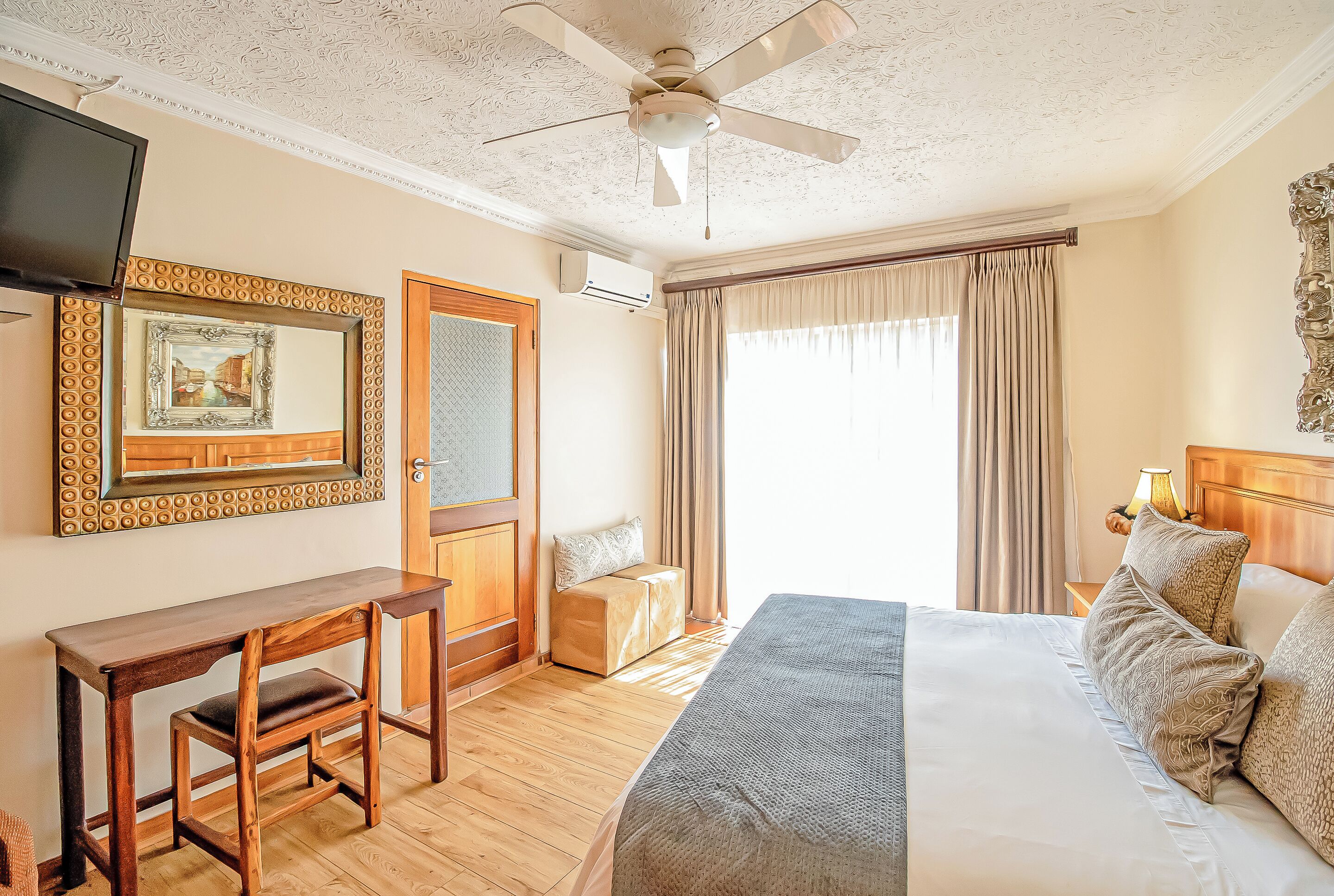 Habitación de hotel con un escritorio de madera, una cama con almohadones estampados, un ventilador de techo y un ventanal con cortinas.