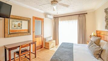 Habitación de hotel con un escritorio de madera, una cama con almohadones estampados, un ventilador de techo y un ventanal con cortinas.