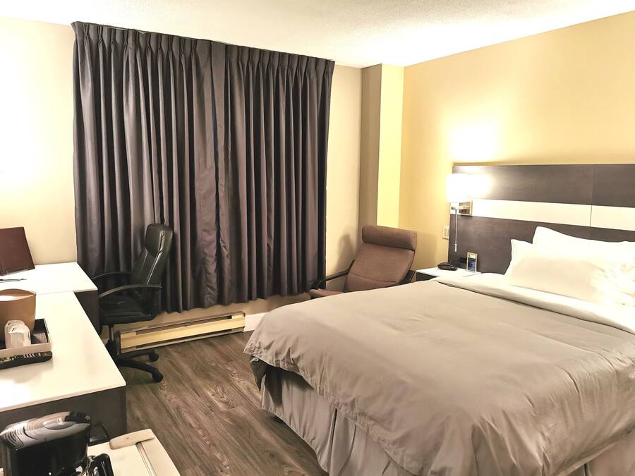 Habitación de hotel con cama, escritorio con computadora portátil, silla, cafetera y ventana grande con cortinas.