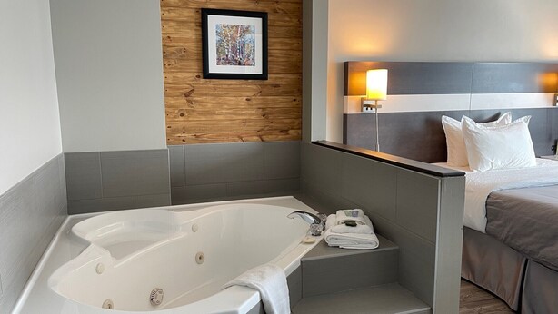 A modern bathroom with a freestanding bathtub, a wooden accent wall, and a bed with a headboard and bedside lamps. - Auberge de la Gare (Gatineau)