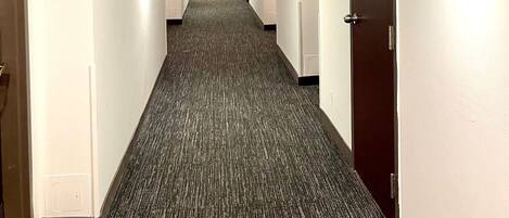 Un pasillo largo y estrecho con pisos alfombrados, vigas de madera en el techo y puertas a ambos lados.