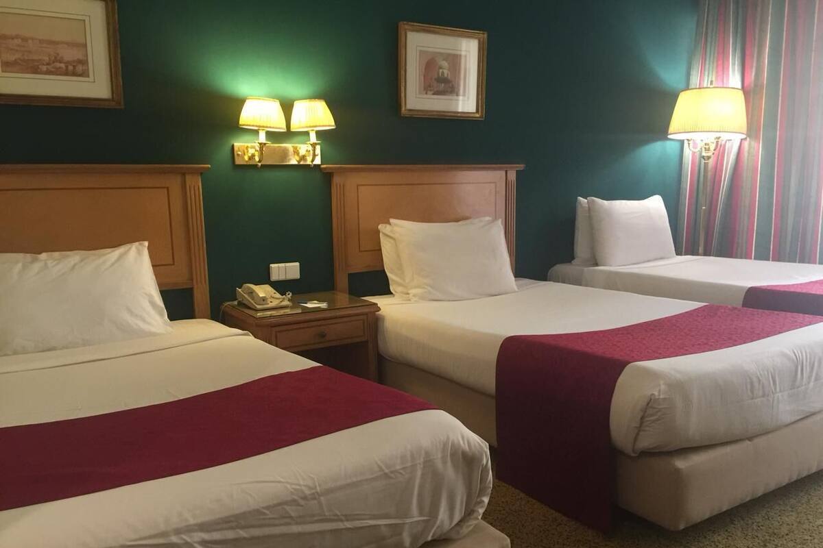 A hotel room with two beds, each with white linens and a red blanket. There are two bedside tables with lamps, a telephone, and a framed picture on the wall.