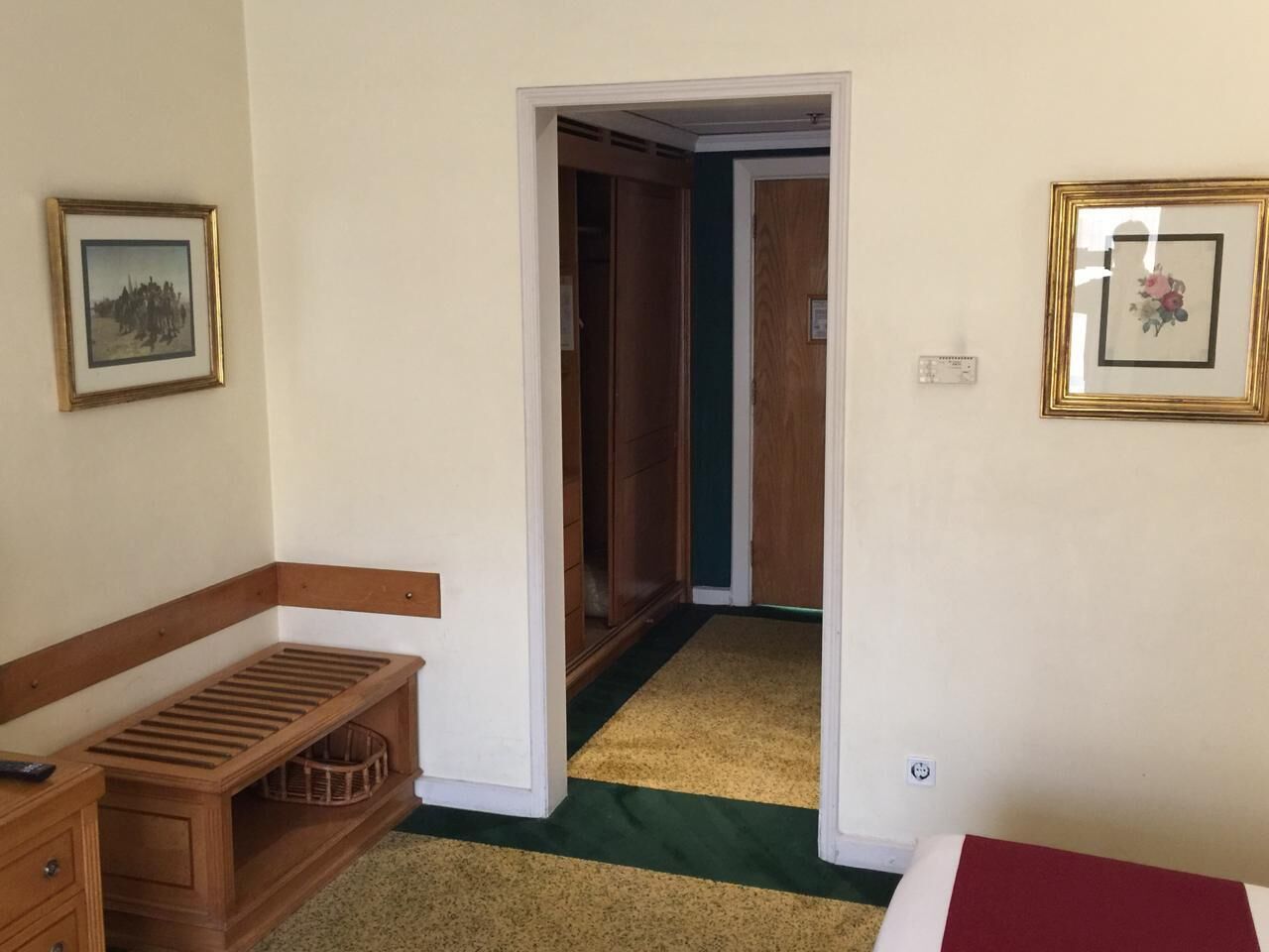 A room with a wooden bench, a small table, and a bed. There is a door leading to another room.