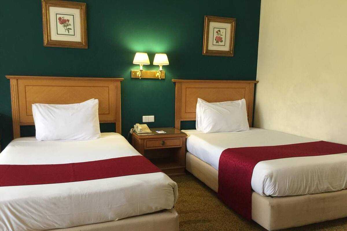 A hotel room with two beds, each with white linens and red blankets, a wooden headboard, a nightstand with a lamp, and framed floral artwork on the walls.
