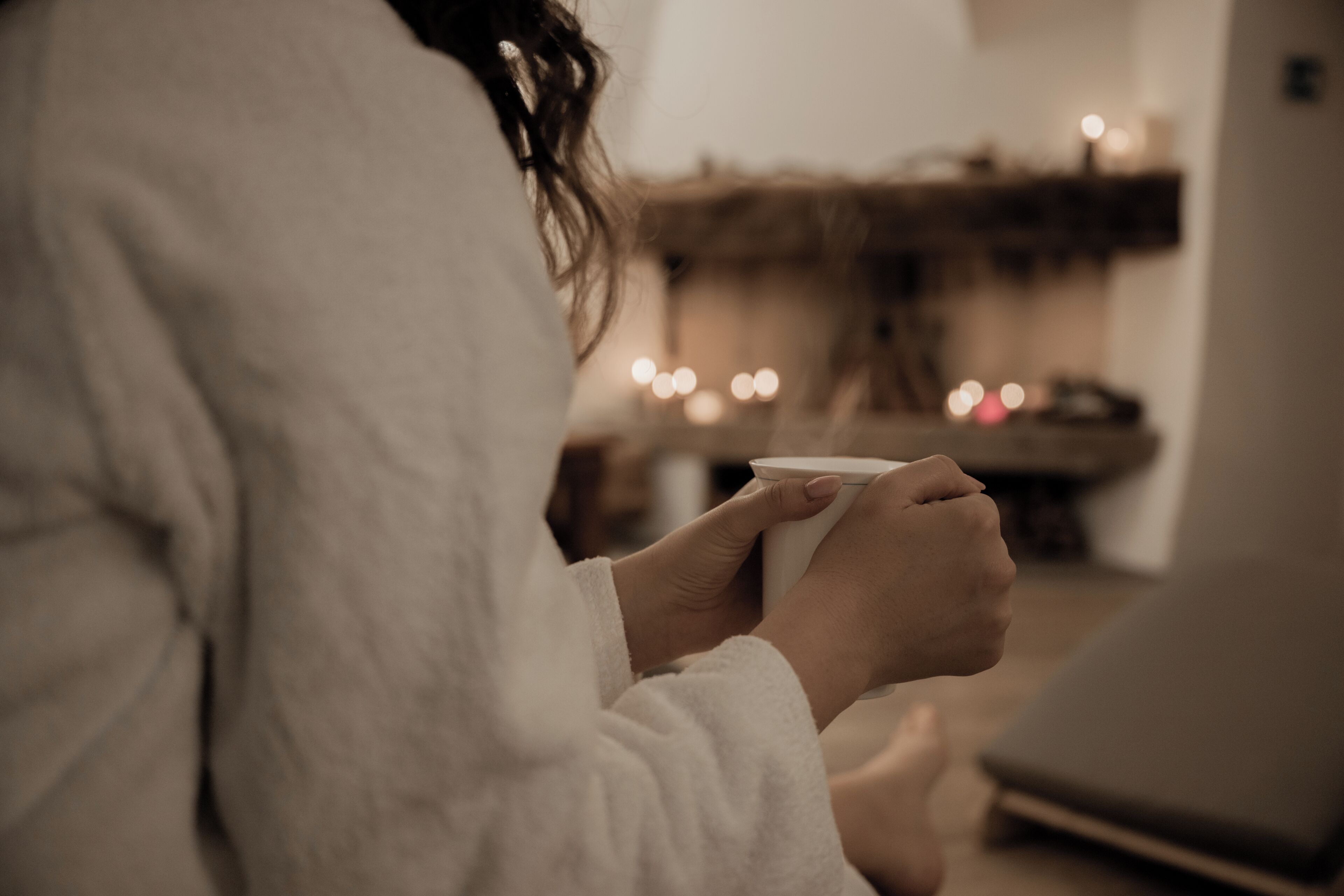 A person in a white robe holding a cup, sitting in a room with a blurred background.