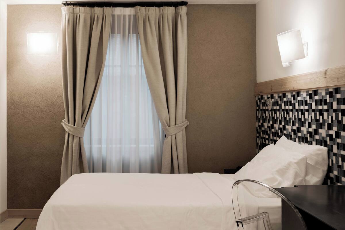 A hotel room with a bed, a bathroom mirror, a tiled wall, and a window with curtains.