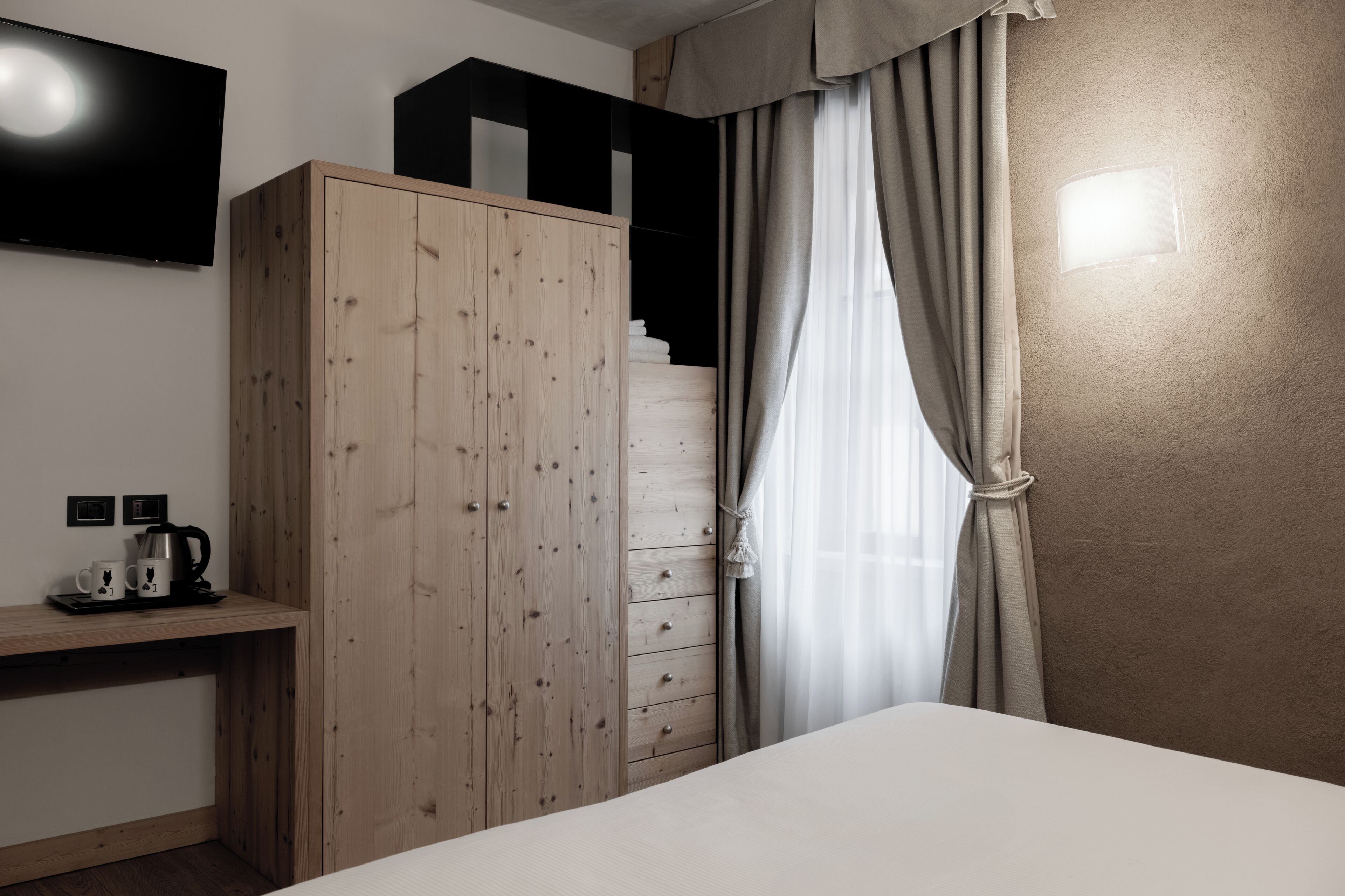 Una habitación de hotel con cama, armario de madera, televisor de pantalla plana, cafetera y ventana con cortinas.